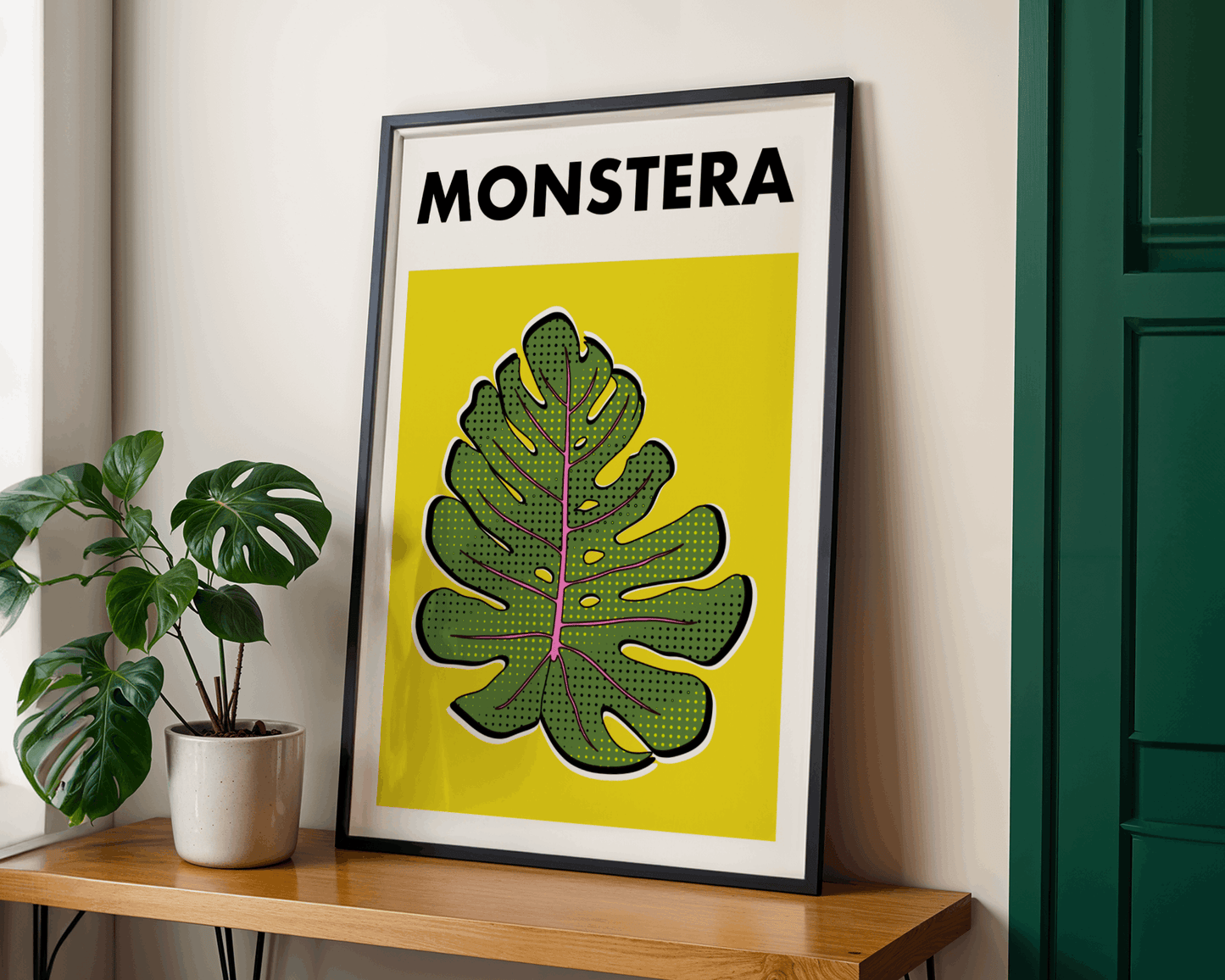 Framed poster with a leaf design and 'MONSTERA' text on a yellow background, placed on a wooden surface next to a potted plant.
