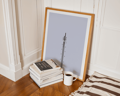 Framed artwork of a skier leaning against a wall next to books and a mug on a wooden floor.