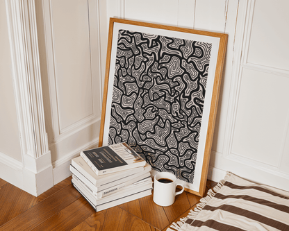 Framed black and white abstract line art leaning against a wall with books and a mug nearby.