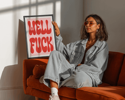 Woman sitting on a couch holding a framed artwork with red text saying WELL FUCK.