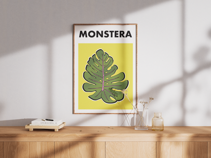 Framed poster with a green monstera leaf design and 'MONSTERA' text on a yellow background, placed on a wooden surface.