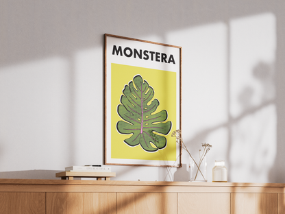 Framed poster with a monstera leaf design and 'MONSTERA' text on a yellow background, placed on a wooden surface.