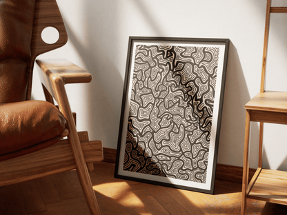 Framed abstract topography artwork on a wooden floor with a brown leather chair and wooden table in the background.